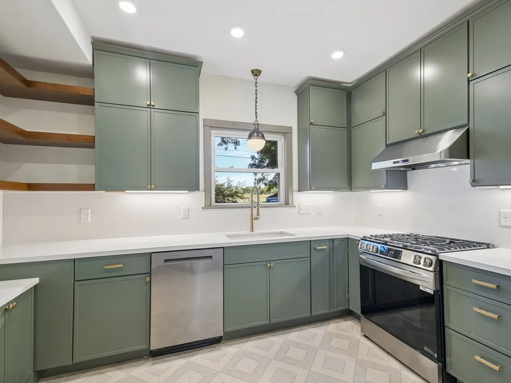 The finished kitchen in the same house — green shaker cabinets, white quartz counters and backsplash, patterned tile floor, stainless appliances and floating wood shelves